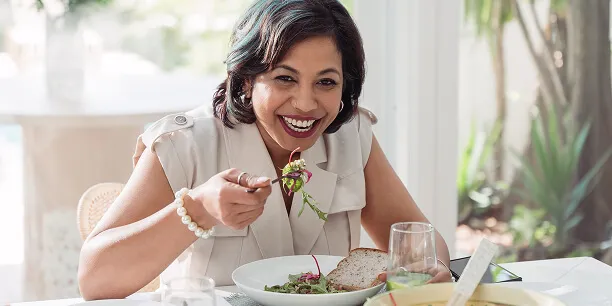 A smiling woman sits at a dining table, holding a forkful of salad and a piece of bread, dining in a bright room with greenery in the background.